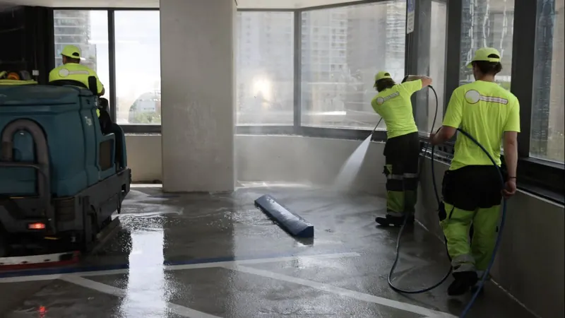 Crew pressure washing a garage wall and deck while a ride-on scrubber cleans the floor